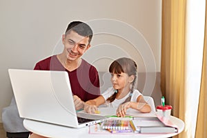 Smiling father and a little daughter doing homework at home, sitting at table in room, looking at laptop screen with smiles,