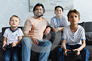 smiling father looking at little sons sitting on sofa and playing video games together