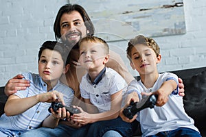 smiling father looking at little sons sitting on sofa and playing video games together