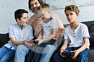 smiling father looking at little sons sitting on sofa and playing video games together