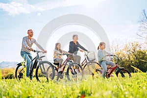 Smiling father and mother with two daughters during summer outdoor bicycle riding. They enjoy togetherness on green high grass