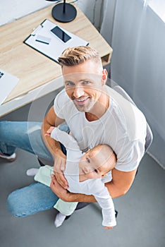 smiling father sitting on chair, holding baby daughter and looking