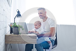 smiling father sitting on chair, holding baby and using laptop