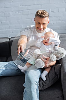 smiling father sitting on couch with baby daughter, holding remote control and watching television