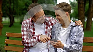 Smiling father and son laughing at funny photo of relative on smartphone, rest