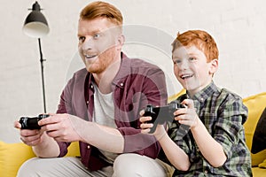 smiling father and son sitting on sofa and playing with joysticks