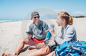 Smiling Father in sunglasses with smartphone sitting with teenager son with backpacks on the sandy seaside beach. Boy drinking wat