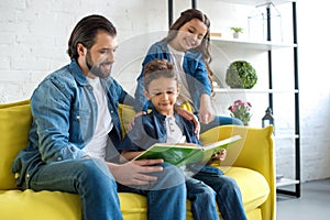 smiling father with two kids reading book together while sitting on couch