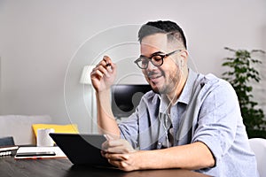 Smiling and feeling positive. Happy Asian man wearing earphones having a video call on a tablet sitting on a desk in the living ro