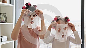 Smiling female applying face mask and curling hair at weekend in bedroom.
