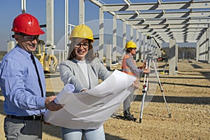 Smiling Female Architect And Supervisor Checking The Blueprint At Future Factory Construction Site