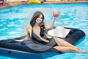 Smiling female in black bikini holding a cocktail sitting on mattress in swimming pool on a blurred background of resort