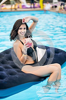 Smiling female in black bikini holding a cocktail sitting on mattress in swimming pool on a blurred background of resort