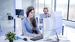 Smiling female customer service consultant with headset using computer and talking with client during phone call.