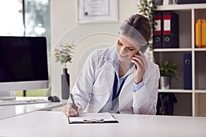 Smiling Female Doctor Or GP Wearing White Coat Sitting At Desk In Office Making Call On Mobile Phone