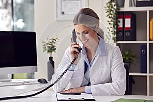 Smiling Female Doctor Or GP Wearing White Coat Sitting At Desk In Office Making Phone Call