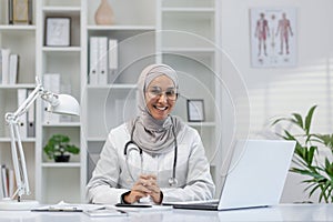 Smiling female doctor in hijab using laptop in clinic office