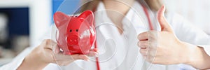 Smiling female doctor holding red pig piggy bank and thumbs up