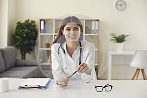 Smiling female doctor looking at camera sits by table. Doctor's office and hospital interior.