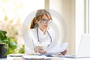 Smiling female doctor sitting at desk and working on laptop photo