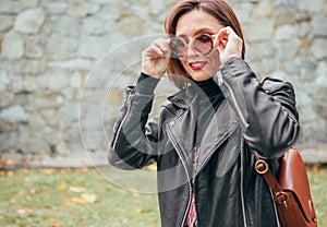 .Smiling female dressed boho fashion style in a black leather biker jacket with brown leather flap bag having an autumn park