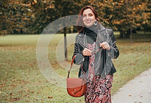 Smiling female dressed boho fashion style colorful long dress with black leather biker jacket with brown leather flap bag having a