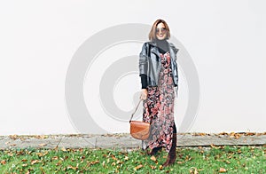 Smiling female dressed boho fashion style colorful long dress with black leather biker jacket with brown leather flap bag posing