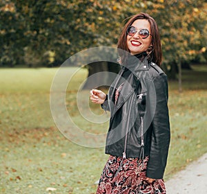 Smiling female dressed boho fashion style colorful long dress with black leather biker jacket in brown sunglasses having an autumn