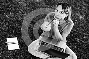 Smiling female entrepreneur sitting on lawn with pc while drinking juice for breakfast - Remote work concept into nature