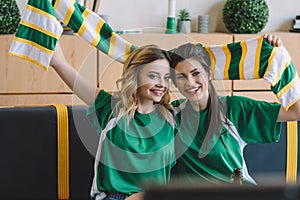 smiling female football fans in green t-shirts and scarf celebrating during watch of soccer match