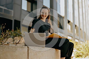 Smiling female freelancer in eyeglasses making notes and browsing laptop outdoors near city building