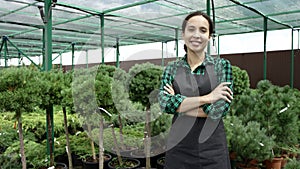 Smiling female gardener standing with arms folded in greenhouse and looking at camera