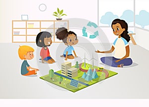 Smiling female kindergarten teacher and children sit on floor and explore toy model with renewable or sustainable energy