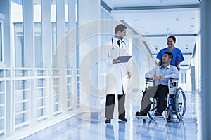 Smiling female nurse pushing and assisting patient in a wheelchair in the hospital, talking to doctor