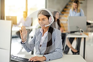 Smiling female operator agent with headset waving hand during video call on computer while having online consultation with