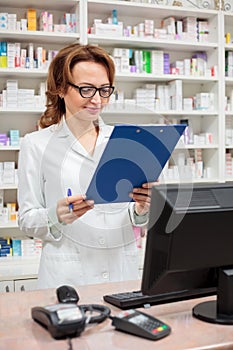 Smiling female pharmacist standing behind the counter in a pharmacy, holding a clipboard and reading documents