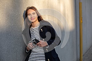 Smiling female photographer in jacket standing in front of wall ready to make new photo. Sun flare