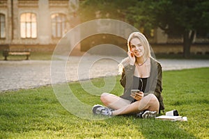 Smiling female student listening to music with a smart phone and headphones sitting on the grass in a campus yard. Cheerful