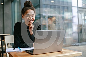 Smiling female student studying on laptop while sitting in cafe and looking on camera