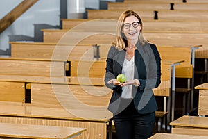 Smiling female teacher in glasses and formal wear holding apple and looking at camera