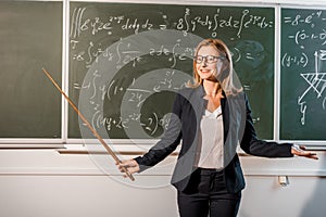 Smiling female teacher with wooden pointer explaining mathematical equations