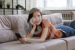 Smiling female teenager lying on belly on couch holding smartphone