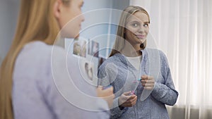 Smiling female teenager putting on lip gloss and looking in the mirror, beauty