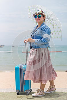 Smiling female tourist holding umbrella and pulling modern blue rolling hard baggage.