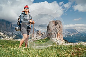 Smiling female trekker with backpack and trekking poles on green mountain hill enjoying picturesque Dolomite Alps. Cinque Torri