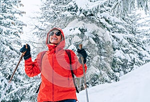 Usmívající se žena trekker oblečená červená bunda ve slunečních brýlích s trekingovými holemi chůzi zasněženým svahem s firtrees pokryté sněhem, nízká