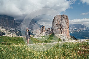 Smiling female trekker walking with backpack and trekking poles by green mountain hill with picturesque Dolomite Alps Cinque Torri