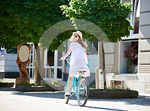 Smiling female in white dress and straw hat looks back to the camera while riding blue bike in paved city center