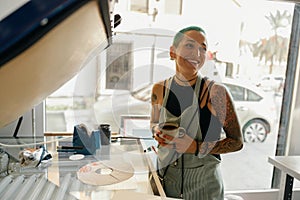 Smiling female worker drink coffee while working with printing machine in a workshop