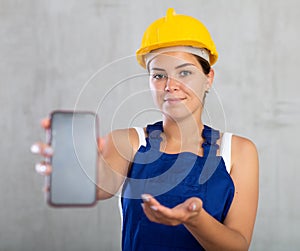 Smiling female worker in work clothes and hardhat demonstrating screen of her phone